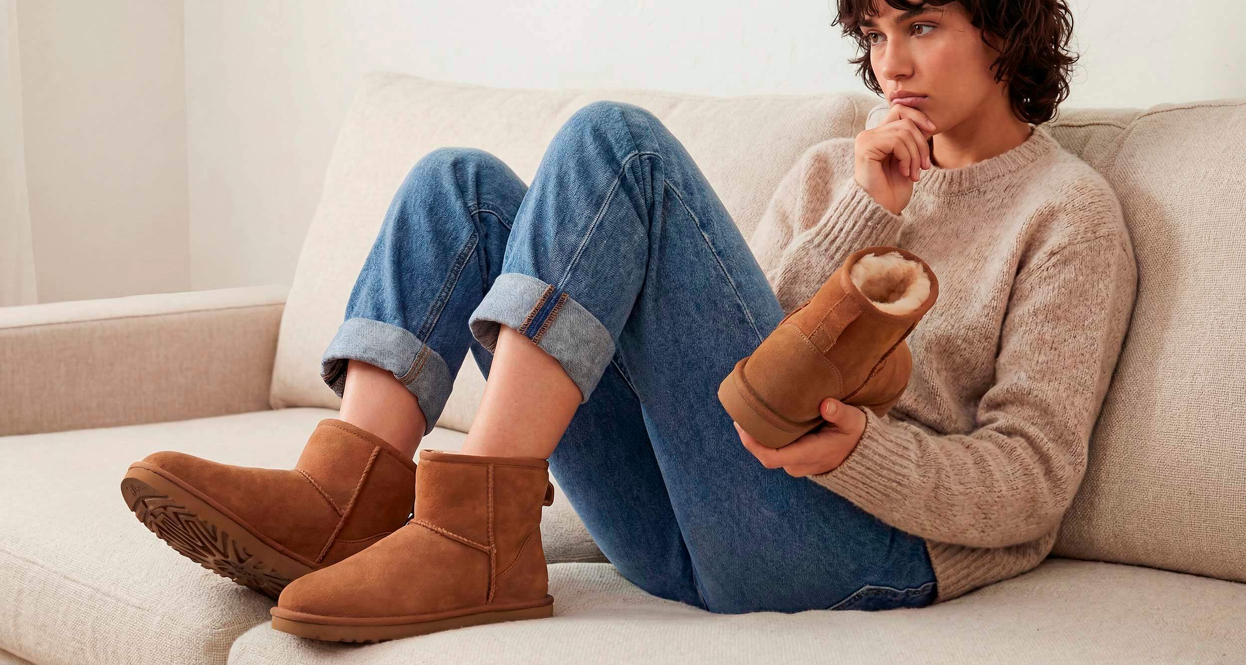 A woman sitting on a sofa trying on a pair of UGG boots, contemplating the fit.